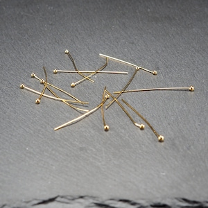 May include: A scattering of gold-toned head pins with round heads, arranged on a dark grey surface.