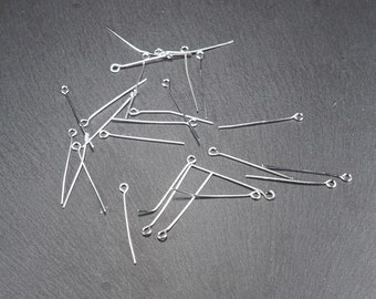 May include: A pile of silver-toned head pins with loops on one end. The pins are scattered on a dark gray surface.