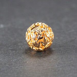 May include: A gold-colored metal bead with a delicate, intricate design of leaves and branches. The bead is hollow and has a round shape.