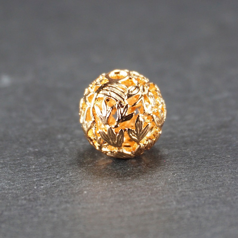 May include: A gold-colored metal bead with a delicate, intricate design of leaves and branches. The bead is hollow and has a round shape.