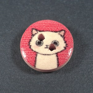 Puede incluir: Un bot&oacute;n de madera blanco con un dise&ntilde;o de gato de dibujos animados. El gato tiene ojos negros y una nariz rosa. El bot&oacute;n est&aacute; sobre un fondo rojo.