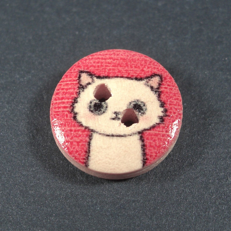 Puede incluir: Un bot&oacute;n de madera blanco con un dise&ntilde;o de gato de dibujos animados. El gato tiene ojos negros y una nariz rosa. El bot&oacute;n est&aacute; sobre un fondo rojo.