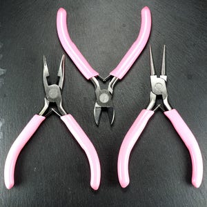 Puede incluir: Tres alicates para joyería con mangos rosas están dispuestos sobre una superficie oscura. Los alicates incluyen unos alicates planos, unos alicates de corte y unos alicates de punta redonda. Las herramientas de metal están diseñadas para manualidades y la fabricación de joyas.