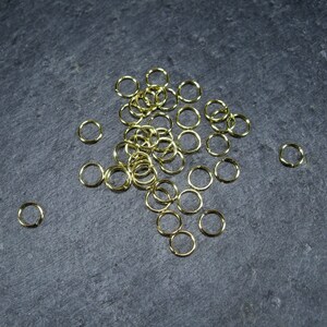 May include: A pile of gold jump rings on a dark grey surface. The jump rings are small and round, and they are perfect for jewellery making.