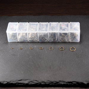 May include: Eight clear plastic containers filled with assorted gold jump rings of different sizes. The containers are arranged in a row on a black surface. Below the containers are seven gold jump rings of varying sizes.