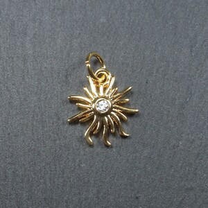Könnte beinhalten: Goldfarbener Sonnenanhänger mit einem klaren Strassstein in der Mitte. Der Anhänger hat eine Öse oben zum Anbringen an eine Halskette oder ein Armband.