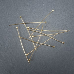 May include: A group of gold-coloured head pins with round heads scattered on a dark grey surface.