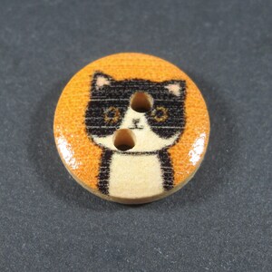 Puede incluir: Un bot&oacute;n de madera con un dise&ntilde;o de gato blanco y negro sobre un fondo naranja. El gato tiene dos ojos negros y una nariz blanca.