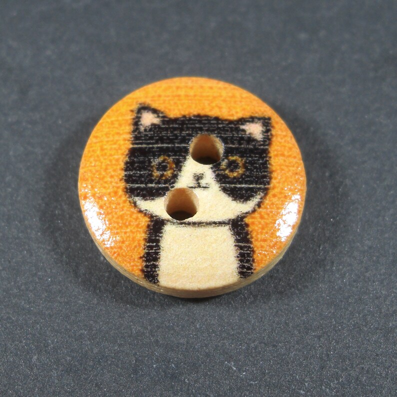 Puede incluir: Un bot&oacute;n de madera con un dise&ntilde;o de gato blanco y negro sobre un fondo naranja. El gato tiene dos ojos negros y una nariz blanca.