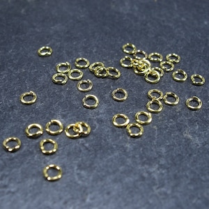May include: A close-up of a pile of gold jump rings on a dark grey surface. The jump rings are small and round, and they are perfect for jewellery making.