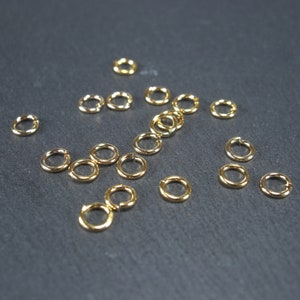 Könnte beinhalten: Eine Sammlung goldener Sprungringe, verstreut auf einer dunkelgrauen Oberfläche. Die Ringe sind klein und rund, mit einer glatten, polierten Oberfläche.