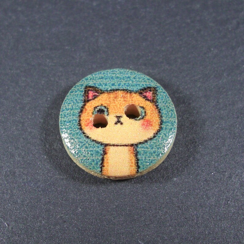 Puede incluir: Un bot&oacute;n de madera con una cara de gato de dibujos animados. El gato tiene pelaje naranja, ojos azules y una nariz rosa. El bot&oacute;n est&aacute; pintado con un fondo azul turquesa.