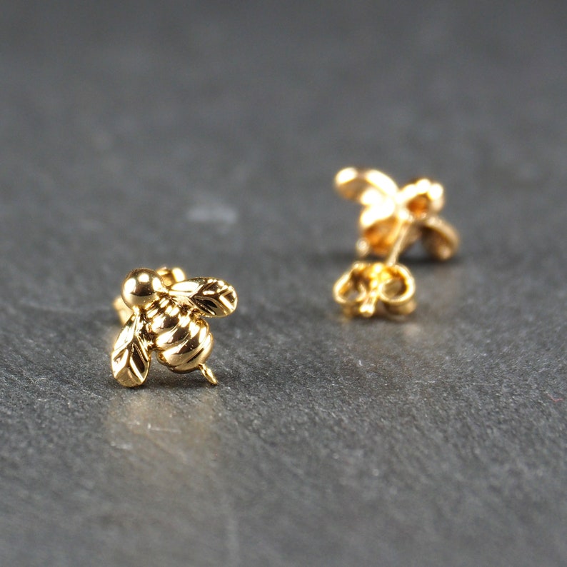 May include: A pair of gold bee stud earrings. The bee is detailed with wings and a textured body.
