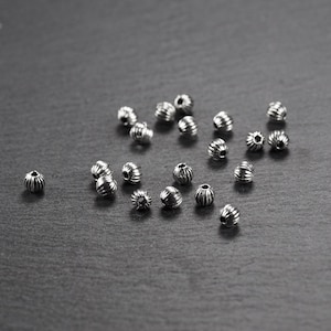 Może przedstawiać: Rozproszenie srebrnych metalowych koralików o fakturowanej, żebrowanej powierzchni. Korale są ułożone na ciemnoszarym tle.