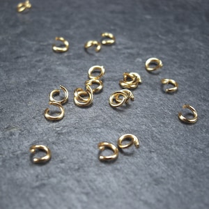 May include: A close-up of a group of gold jump rings scattered on a dark grey surface. The jump rings are small and circular, with a smooth, shiny finish.