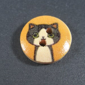 Puede incluir: Un bot&oacute;n redondo de madera con una ilustraci&oacute;n de un gato blanco y negro sobre un fondo amarillo. El gato tiene ojos verdes y una nariz blanca.
