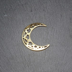 Puede incluir: Un amuleto de media luna dorado con un diseño decorativo e intrincado. El amuleto está hecho de metal y tiene un patrón recortado.