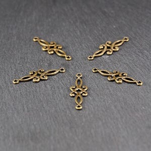 Puede incluir: Cinco adornos decorativos de metal dorado con intrincados dise&ntilde;os de volutas. Los adornos tienen tres agujeros cada uno, lo que sugiere que est&aacute;n destinados a ser utilizados en la fabricaci&oacute;n de joyas u otros proyectos de artesan&iacute;a.