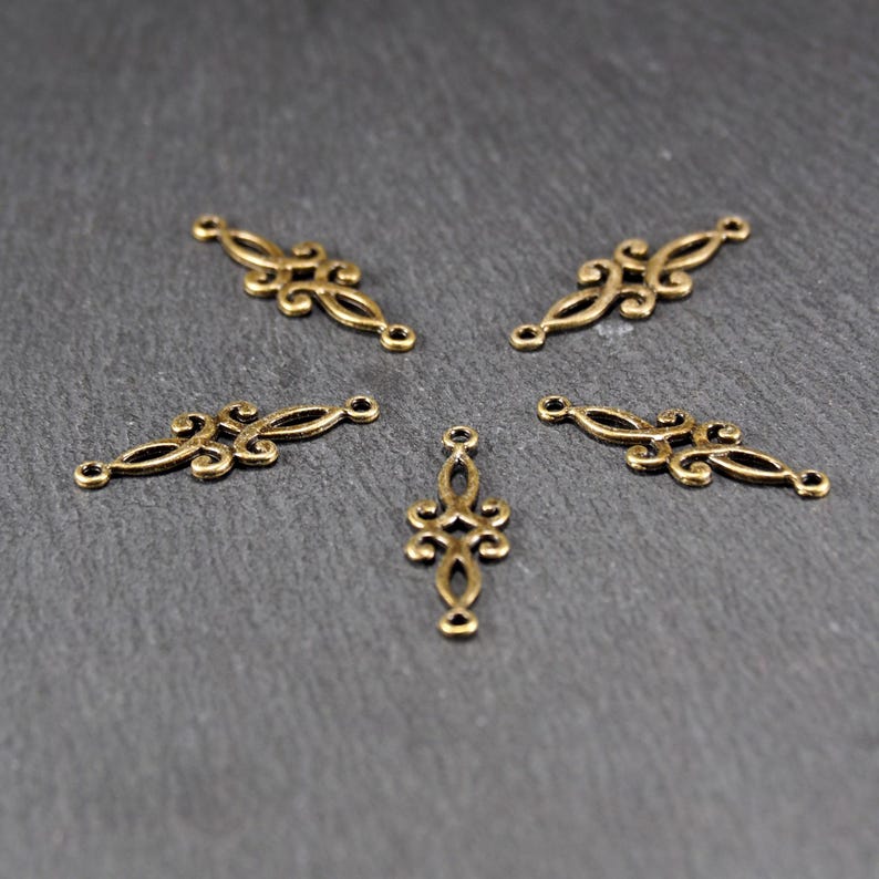 Puede incluir: Cinco adornos decorativos de metal dorado con intrincados dise&ntilde;os de volutas. Los adornos tienen tres agujeros cada uno, lo que sugiere que est&aacute;n destinados a ser utilizados en la fabricaci&oacute;n de joyas u otros proyectos de artesan&iacute;a.