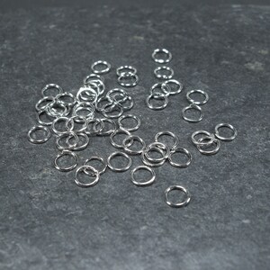 May include: A scattering of small, shiny silver jump rings on a dark grey surface. The circular rings are made of metal, suitable for jewellery making or crafts. The lighting accentuates their reflective nature.
