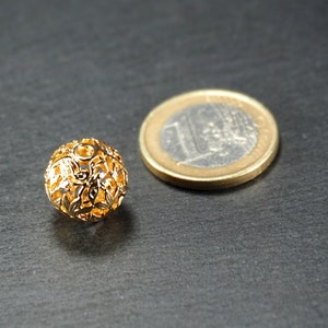 May include: A gold-colored metal bead with a decorative, intricate design. The bead is round and has a hollow center. It is sitting on a dark gray surface next to a euro coin.
