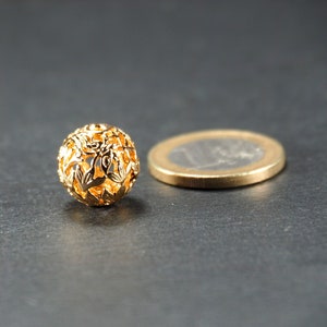 May include: A gold-colored metal bead with a delicate, intricate design. The bead is round and has a hollow center, creating a lacy effect.