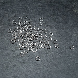 May include: A scattering of small, shiny, silver jump rings on a dark grey surface. These circular metal rings are reflective, likely used for jewellery making or crafts. Each ring is a small, closed circle.