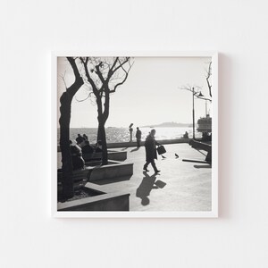 May include: Black and white photograph of people sitting on benches along a waterfront with a ferry in the distance.