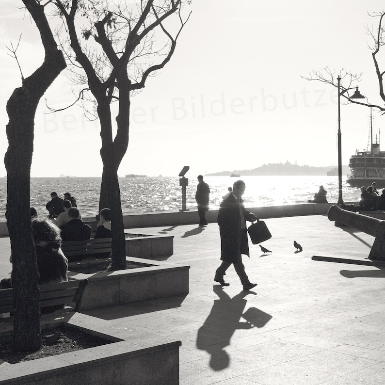 May include: A black and white photo of a man walking on a sidewalk near a body of water. There are trees and other people in the background.