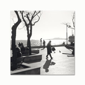 May include: A black and white photo of people sitting on benches and walking along a waterfront with a ferry in the distance.