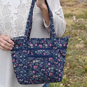 Op de afbeelding: Marineblau gewatteerde tote bag met een bloemenpatroon in roze, geel en groen. De tas heeft twee buitenzakken en stevige handvatten. De tas is ontworpen met een ruitpatroon.