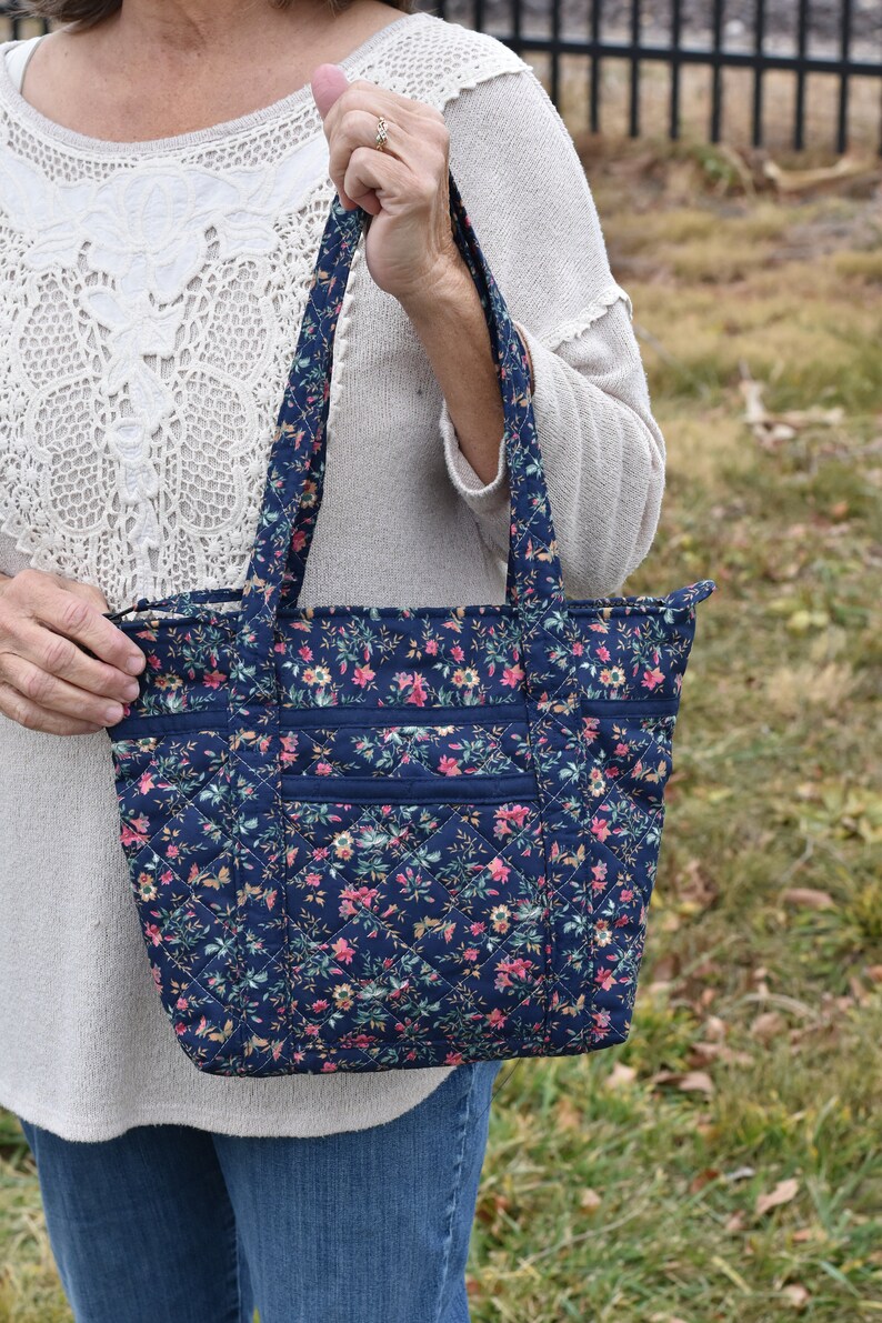 Op de afbeelding: Marineblau gewatteerde tote bag met een bloemenpatroon in roze, geel en groen. De tas heeft twee buitenzakken en stevige handvatten. De tas is ontworpen met een ruitpatroon.