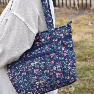 Op de afbeelding: Een marineblauwe gewatteerde tote bag met een bloemenpatroon in roze, geel en groen. De tas heeft een ruitpatroon, twee buitenvakken en bijpassende bandjes. De tas is ontworpen voor het dragen van spullen.