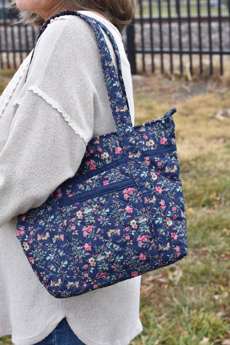 Op de afbeelding: Een marineblauwe gewatteerde tote bag met een bloemenpatroon in roze, geel en groen. De tas heeft een ruitpatroon, twee buitenvakken en bijpassende bandjes. De tas is ontworpen voor het dragen van spullen.