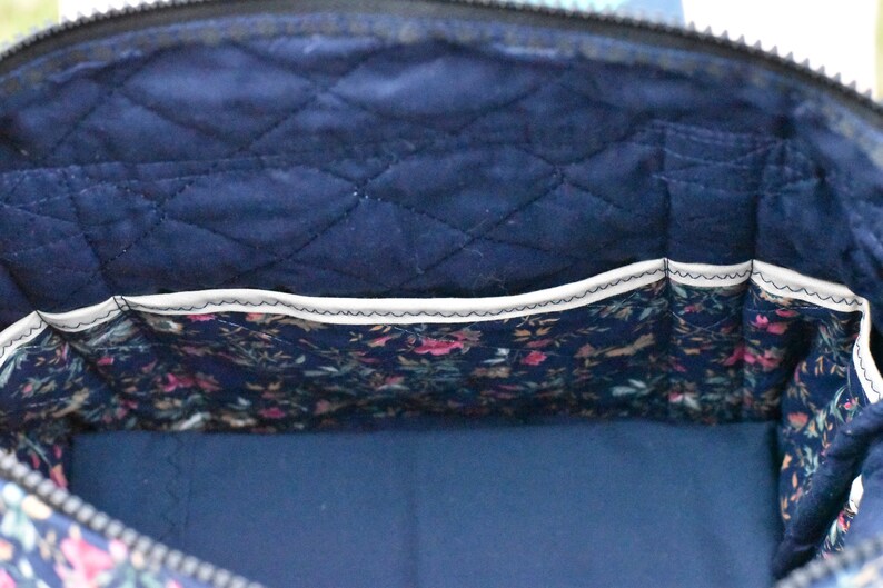Op de afbeelding: Binnenaanzicht van een marineblauwe gewatteerde tas met een bloemenprintzak. De binnenkant van de tas heeft een ruitvormig stikselpatroon. De zak is gevoerd met een bloemenstof in roze, groene en blauwe tinten. De tas heeft een rits.