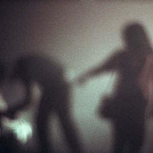 Op de afbeelding: Twee wazige silhouetten van mensen die voor een muur staan. De persoon rechts kijkt naar de camera en de persoon links kijkt weg van de camera.