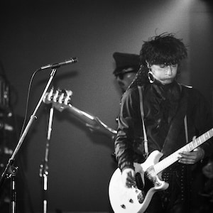 Peut inclure: Une photo en noir et blanc d'un musicien jouant de la guitare électrique blanche avec une sangle noire. Le musicien porte une veste noire et a les cheveux longs et foncés. Le musicien se tient sur scène avec un pied de micro et un amplificateur de guitare en arrière-plan.