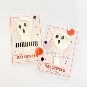 May include: Two off-white ghost-shaped balloons attached to rectangular cards with black and white tape. The cards feature the text "happy HALLOWEEN" and "FROM: Avery" with colorful confetti accents. Each balloon is sealed in a clear plastic sleeve.