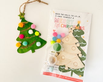 May include: A green wooden Christmas tree ornament with colorful pom-poms and glitter accents, next to a kit with a wooden tree, paint, and the text "MERRY little CHRISTMAS". The ornament has a twine loop for hanging.