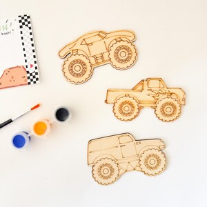 May include: Three unfinished wooden monster truck shapes with paintbrushes and paint pots in blue, orange, and black. The trucks are ready to be painted.