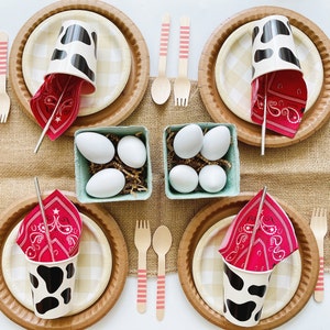 May include: A table setting for a farm-themed party with brown and white checkered paper plates, red and white bandana napkins, black and white cow-print cups, wooden spoons and forks, and a small green container filled with white eggs.