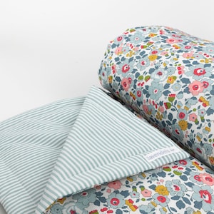 Könnte beinhalten: Ein Kissen- und Deckenset mit floralem Muster. Das zylindrische Kissen ist mit einem blauen, rosa und gelben Blumenmuster versehen. Die Decke hat auf der einen Seite ein Streifenmuster und auf der anderen Seite ein Blumenmuster. Die Decke ist gefaltet.