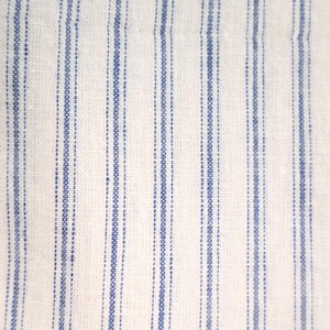 Può includere: Primo piano di un tessuto bianco con strisce blu verticali. Le strisce sono uniformemente distanziate e sembrano essere ricamate o tessute nel tessuto. La trama del tessuto è visibile, suggerendo una fibra naturale come il cotone o il lino.