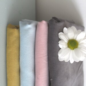 Op de afbeelding: Vier rollen stof in verschillende kleuren: mosterdgeel, lichtblauw, roze en grijs. De stof is geribbeld en heeft een corduroy textuur. Een witte madeliefje ligt bovenop de grijze stof.