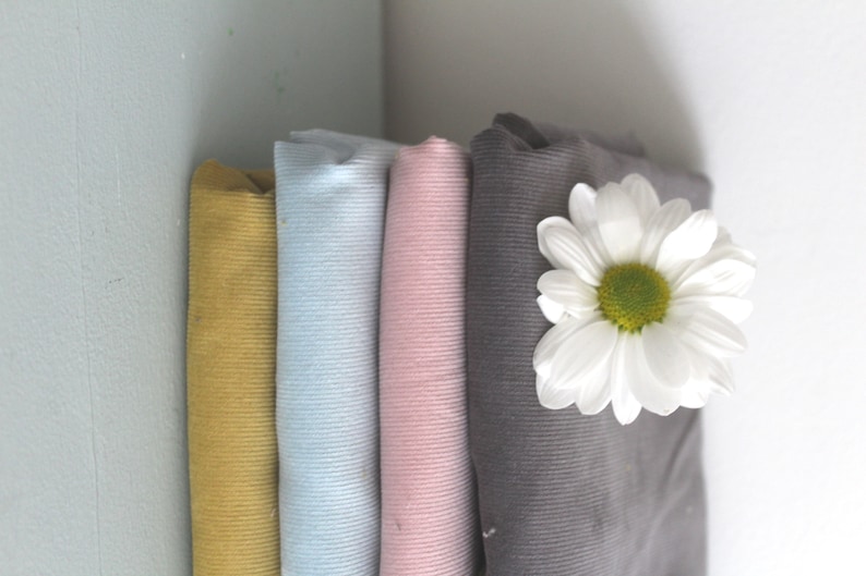 Op de afbeelding: Vier rollen stof in verschillende kleuren: mosterdgeel, lichtblauw, roze en grijs. De stof is geribbeld en heeft een corduroy textuur. Een witte madeliefje ligt bovenop de grijze stof.