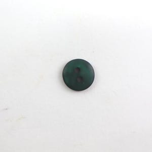Könnte beinhalten: Ein dunkelgrüner, runder Knopf mit zwei Löchern. Der Knopf besteht aus einem festen Material und hat eine leicht strukturierte Oberfläche. Geeignet für Nähprojekte und Kleidung.
