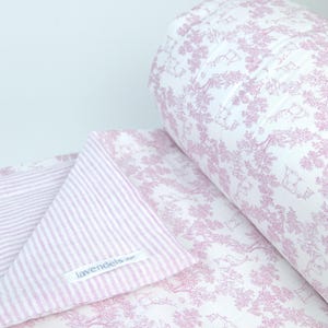 Può includere: Una trapunta bianca con un motivo toile rosa ripetuto di alberi e animali. La trapunta è piegata per rivelare un lato rovescio con un motivo a righe rosa e bianche. Un'etichetta in tessuto bianco con il testo lavendeis è cucita sul tessuto a righe.