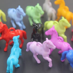 May include: A collection of small, plastic horse-shaped charms in various solid colours including red, blue, yellow, green, pink, purple, brown, black, and white. Each horse figure features a small loop on its back for attachment purposes.