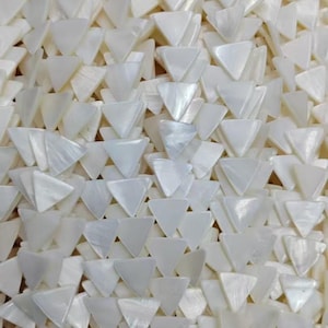 Puede incluir: Primer plano de una colección de objetos blancos en forma de triángulo. Los objetos están dispuestos en un patrón en capas, creando una superficie texturizada. Los triángulos tienen un brillo nacarado, que refleja la luz.