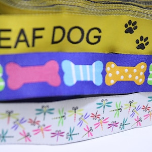 May include: Three yellow and black ribbons with the text "DEAF DOG" printed on them. The ribbons are layered on top of each other. The bottom ribbon has a white background with a repeating pattern of colourful dragonflies. The middle ribbon has a blue background with a repeating pattern of colourful bone-shaped designs.